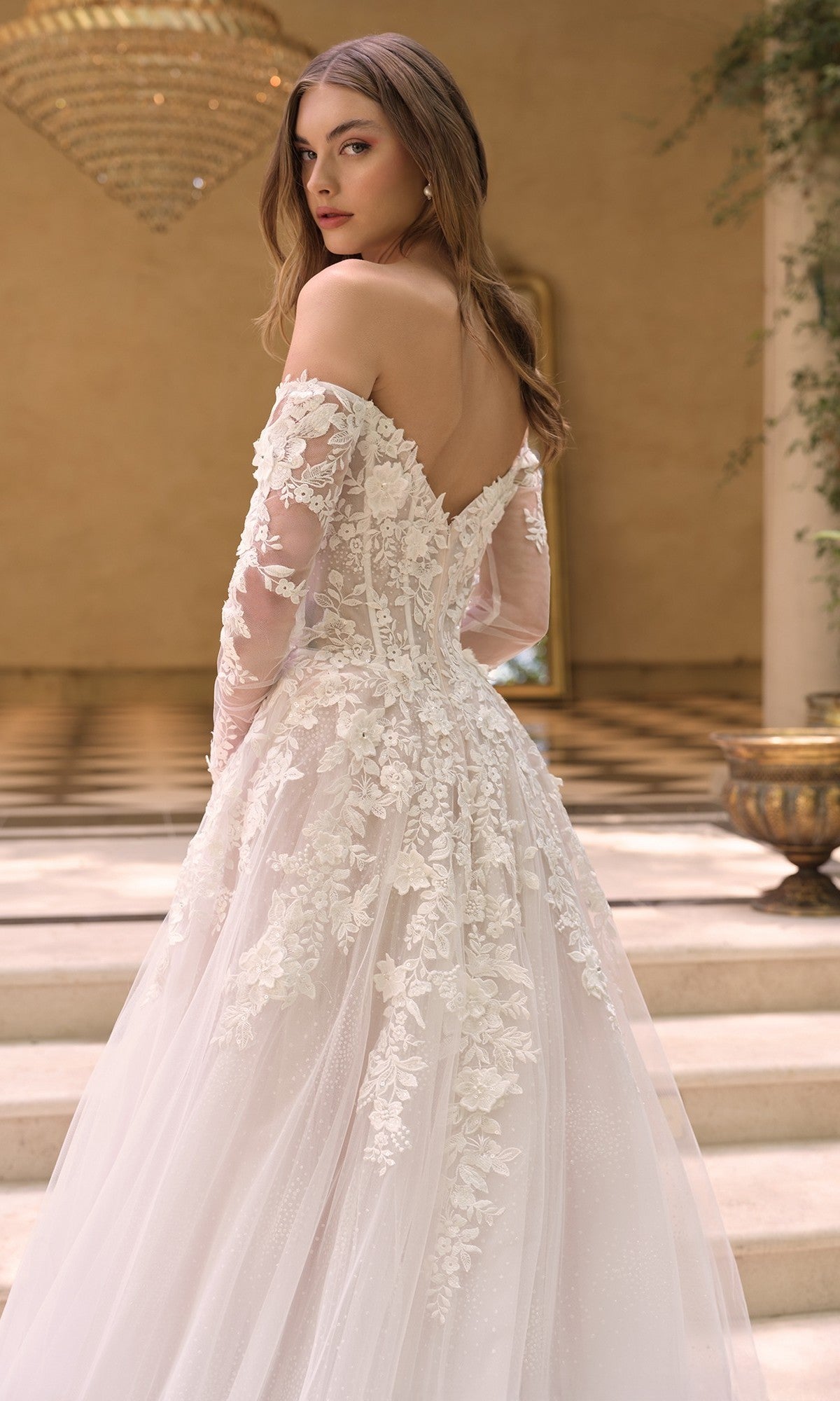 A woman stands on steps in an elegant indoor setting, wearing the White Lace Wedding Dress: Andrea and Leo WL067—an off-the-shoulder, long-sleeved gown with a delicate corset bodice and 3D floral lace appliqué.